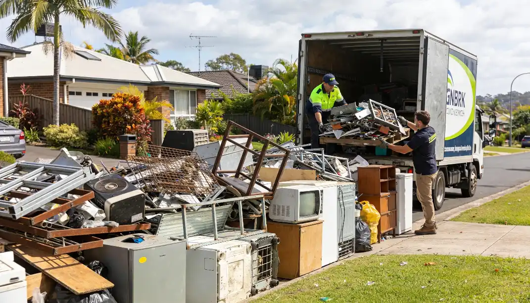 Two workers in hi-vis vests loading large amounts of bulk waste including old televisions furniture and scrap metal into a council collection truck parked on a Brisbane suburban street.