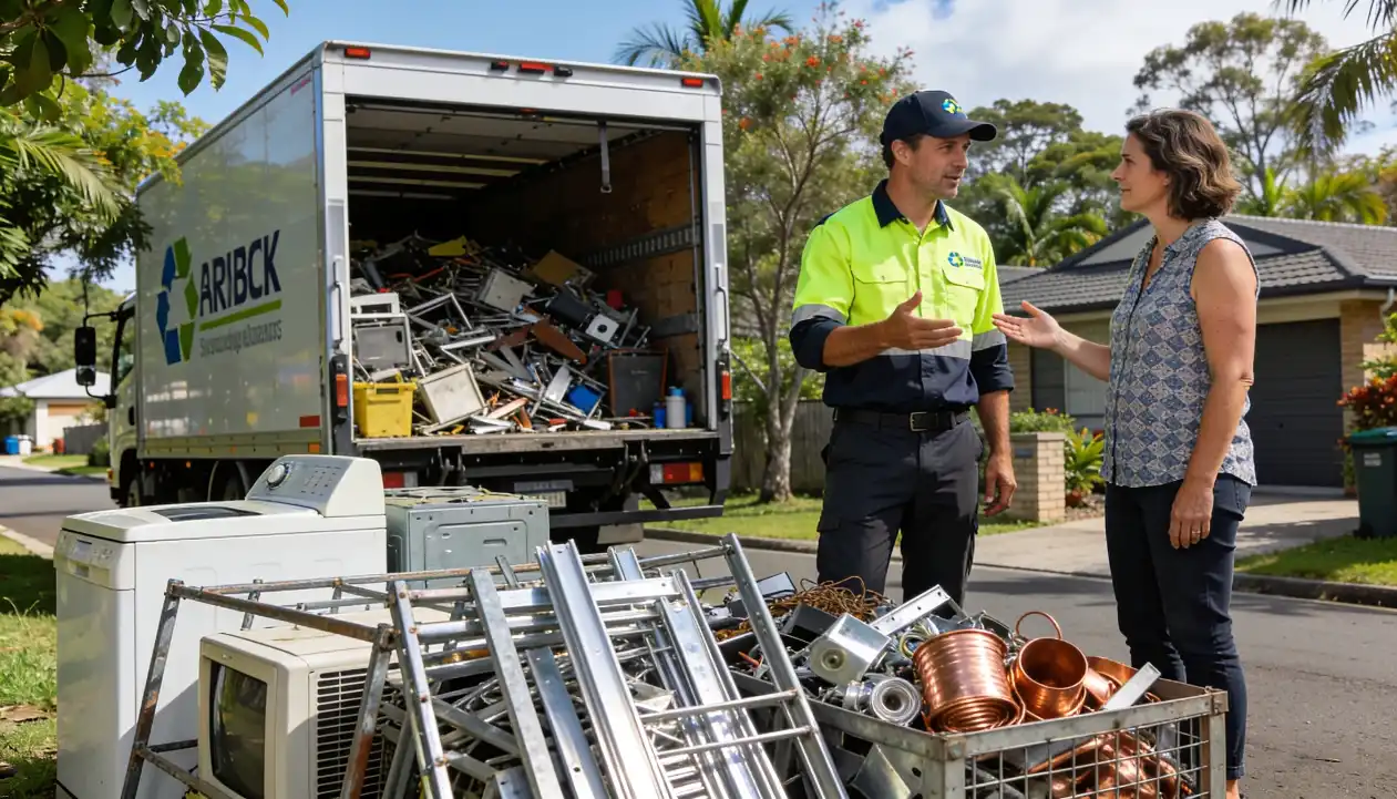 A recycling service professional in a hi-vis vest discussing responsible scrap disposal with a female homeowner beside scrap metal, copper pipes and old appliances.